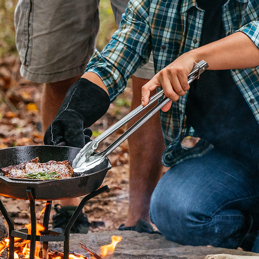 Someone using tongs to flip steak while cooking in a skillet over campfire.