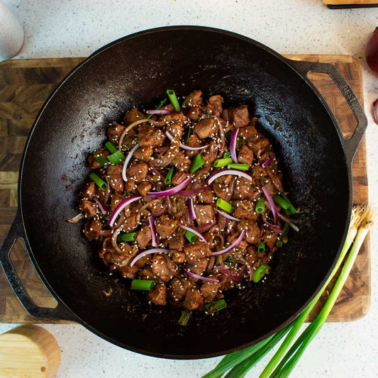 Fried beef dish with onions and green onions in a cast iron wok on a wooden cutting board.