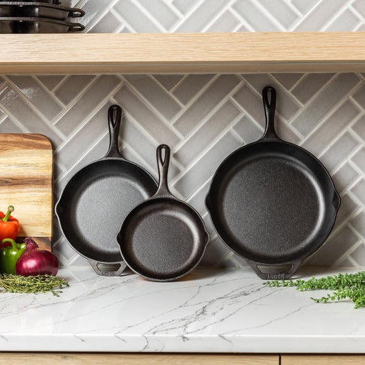 Set of cast iron skillets on a kitchen counter with vegetables and a cutting board.
