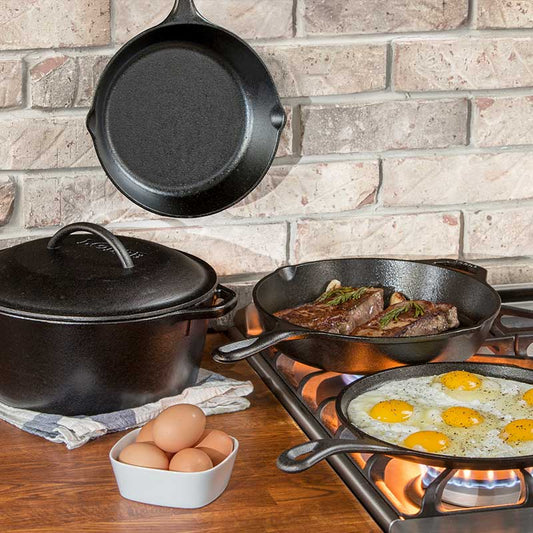 A set of black cast iron cookware including a pot, pan, grill pan, sauté pan, and a skillet, displayed on a stovetop with food being cooked in the pans and a seasoned cast iron label visible.