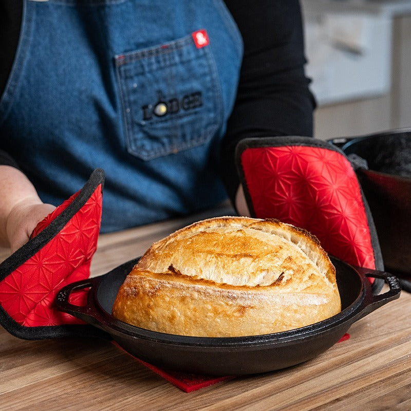 Freshly baked bread in double dutch oven, being placed on counter using silicone-and-fabric potholders.