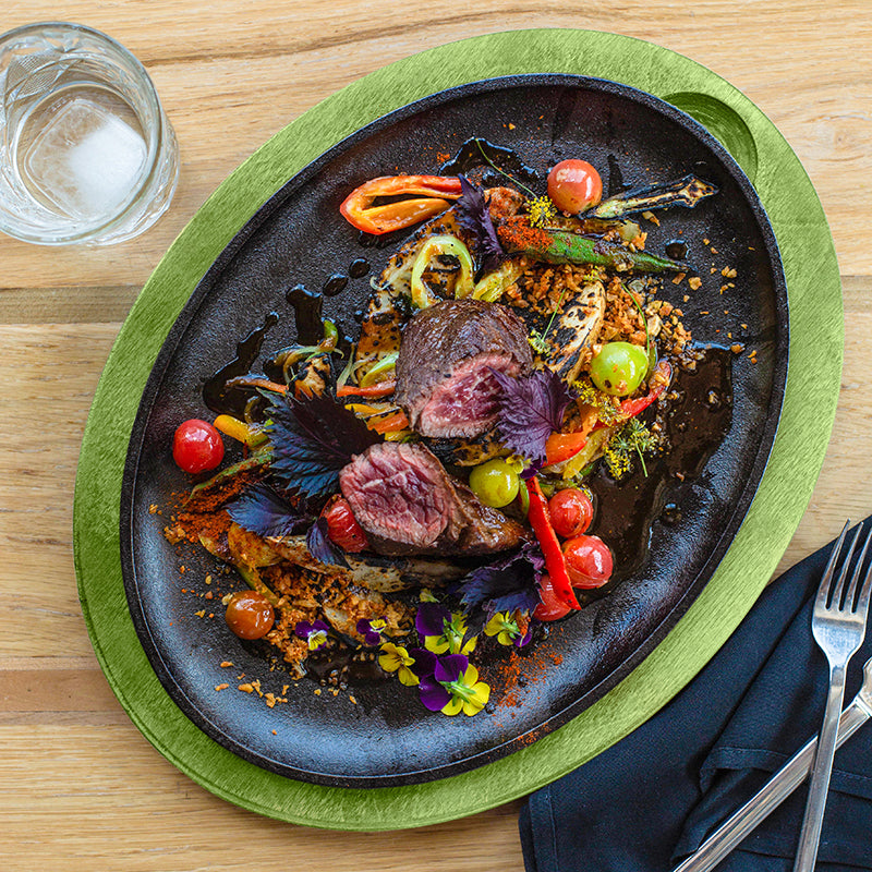 Delicate dish of beef with vegetables on a cast iron serving griddle with a green underliner, served on a kitchen table.