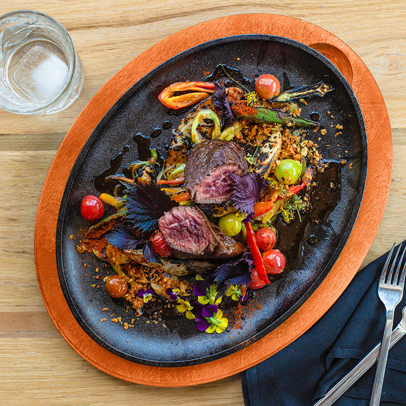 Delicate dish of beef with vegetables on a cast iron serving griddle with an orange underliner, served on a kitchen table.