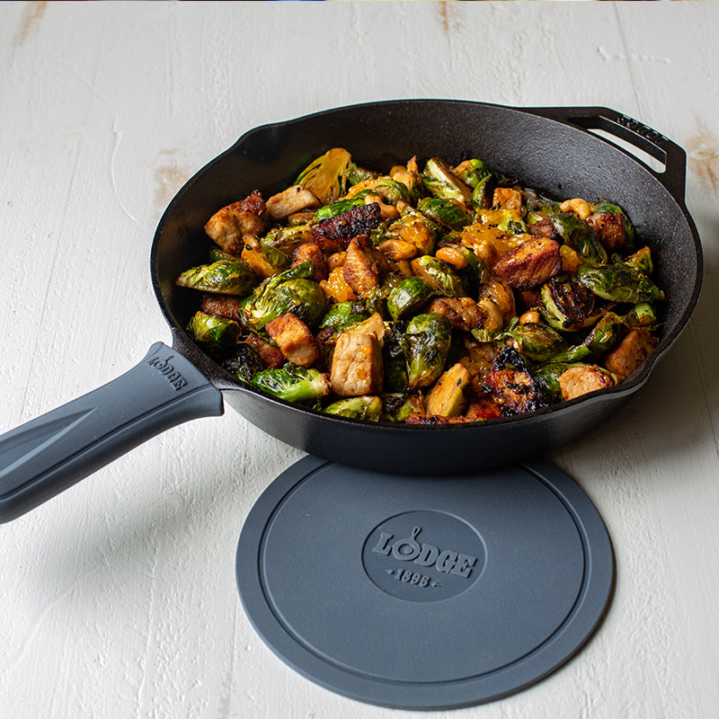 Grey silicone handle holder on skillet with freshly made chicken and Brussel sprouts next to Lodge deluxe silicone trivet.