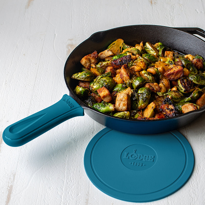 Blue silicone handle holder on skillet with freshly made chicken and Brussel sprouts next to Lodge deluxe silicone trivet.