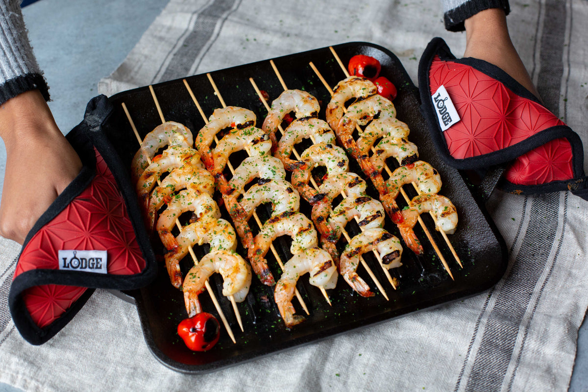 Someone using handle holders to carry our grill pan after cooking shrimp skewers.