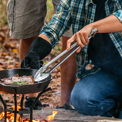 Someone using tongs to flip steak while cooking in a skillet over campfire.