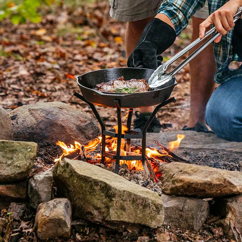 Someone cooking steak with skillet using the stand over a fire.