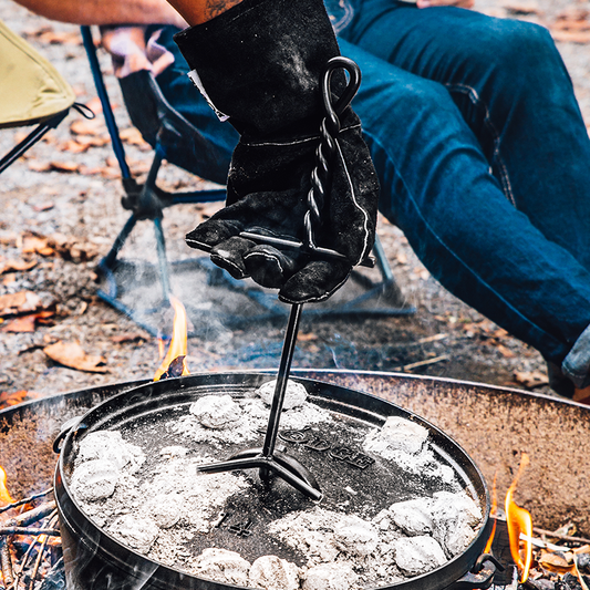 A person is using a black lid lifter to lift the lid of a camp dutch oven placed on a campfire.