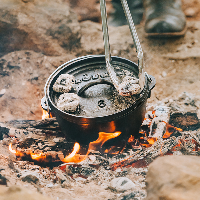 Someone placing coals on lid while dutch is cooking on a fire.