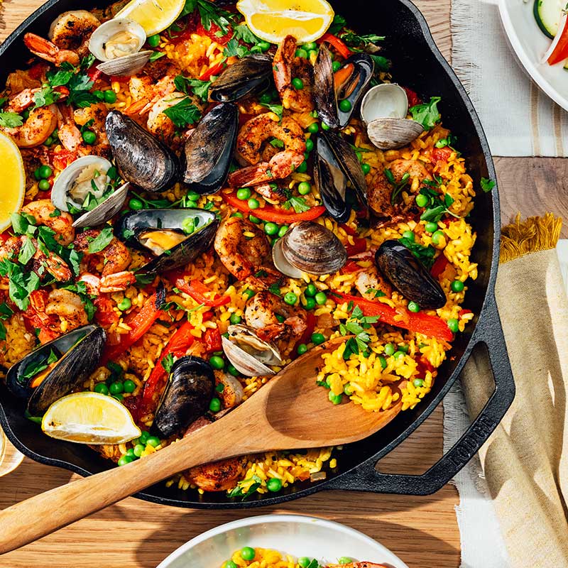A 14 inch dual handle cast iron skillet with a colorful dish of rice topped with seafood and a sprig of parsley, placed on a stovetop with a wooden handle and a lemon slice to the side.