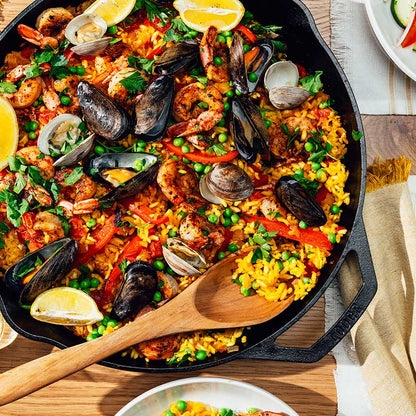 A 14 inch dual handle cast iron skillet with a colorful dish of rice topped with seafood and a sprig of parsley, placed on a stovetop with a wooden handle and a lemon slice to the side.
