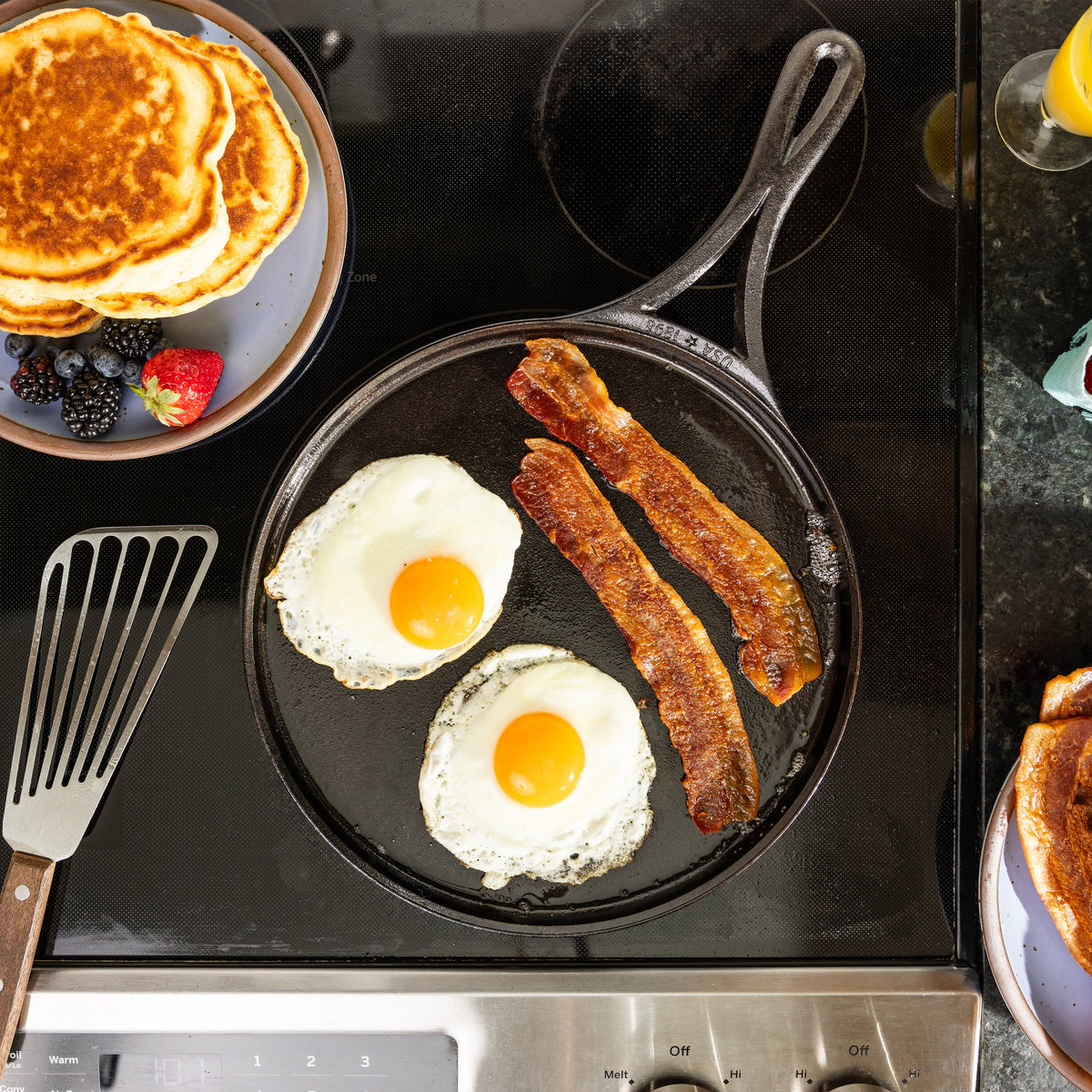 Overhead view of a black cast iron skillet on a black stovetop, cooking two sunny eggs and two strips of bacon. A plate of pancakes, a spatula, and part of a drink are visible around the stove.