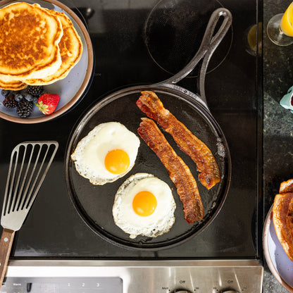 Overhead view of a black cast iron skillet on a black stovetop, cooking two sunny eggs and two strips of bacon. A plate of pancakes, a spatula, and part of a drink are visible around the stove.