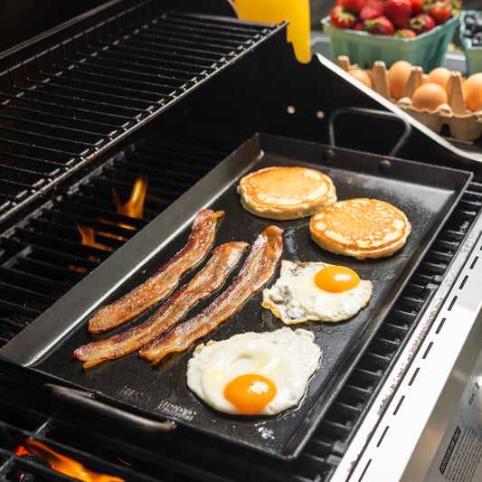 A black flat-top carbon steel griddle cooking pancakes, bacon, eggs, and a sunny-side-up egg on a grill, with a carton of eggs and strawberries visible in the background.