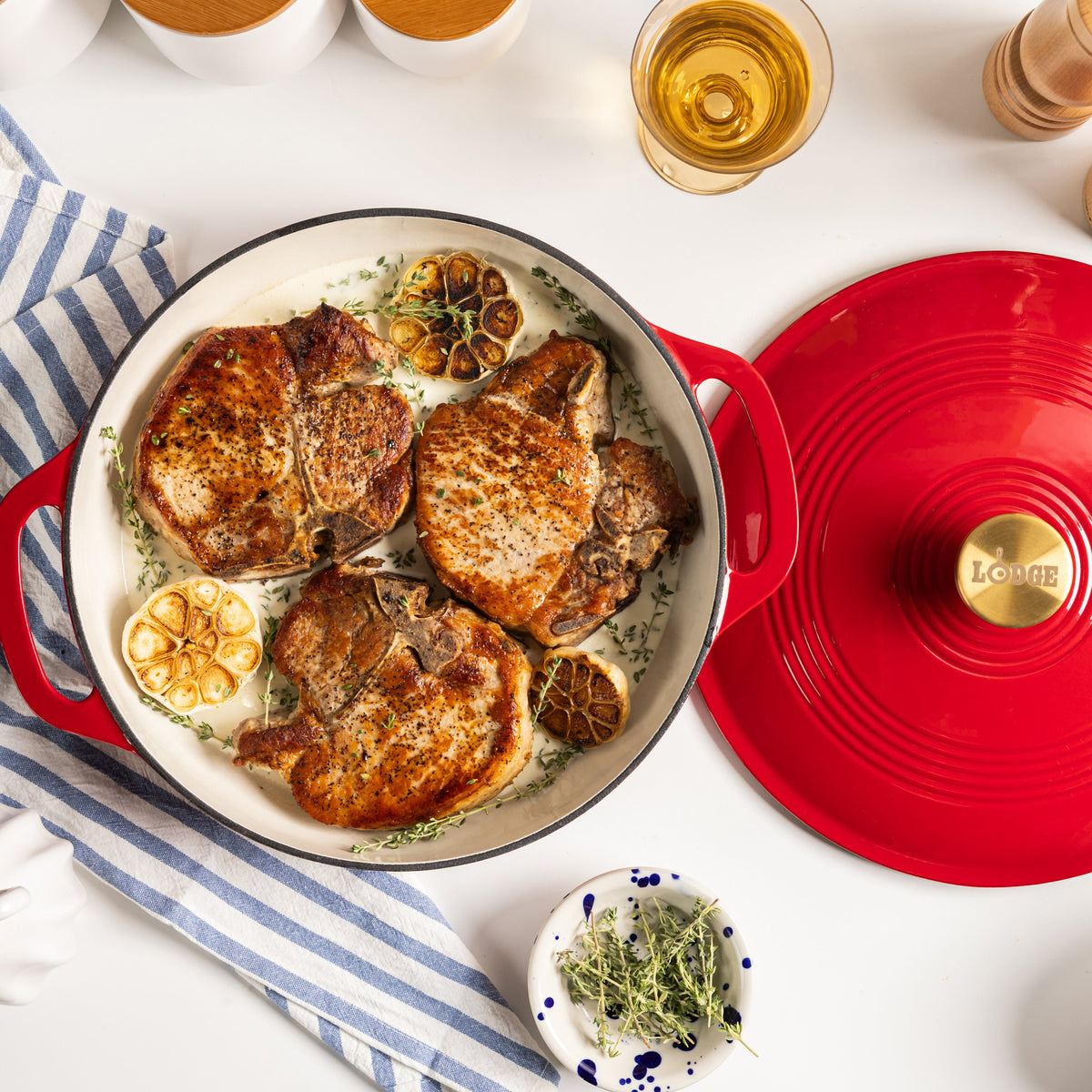 A red enameled cast iron braiser with a matching lid, shown on a white tabletop with cooked pork chops, lemon slices, herbs, and a striped cloth. The lid has a gold-tone knob with the Lodge brand name visible.
