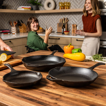Three cast iron skillets on a wooden countertop with two women in a kitchen.