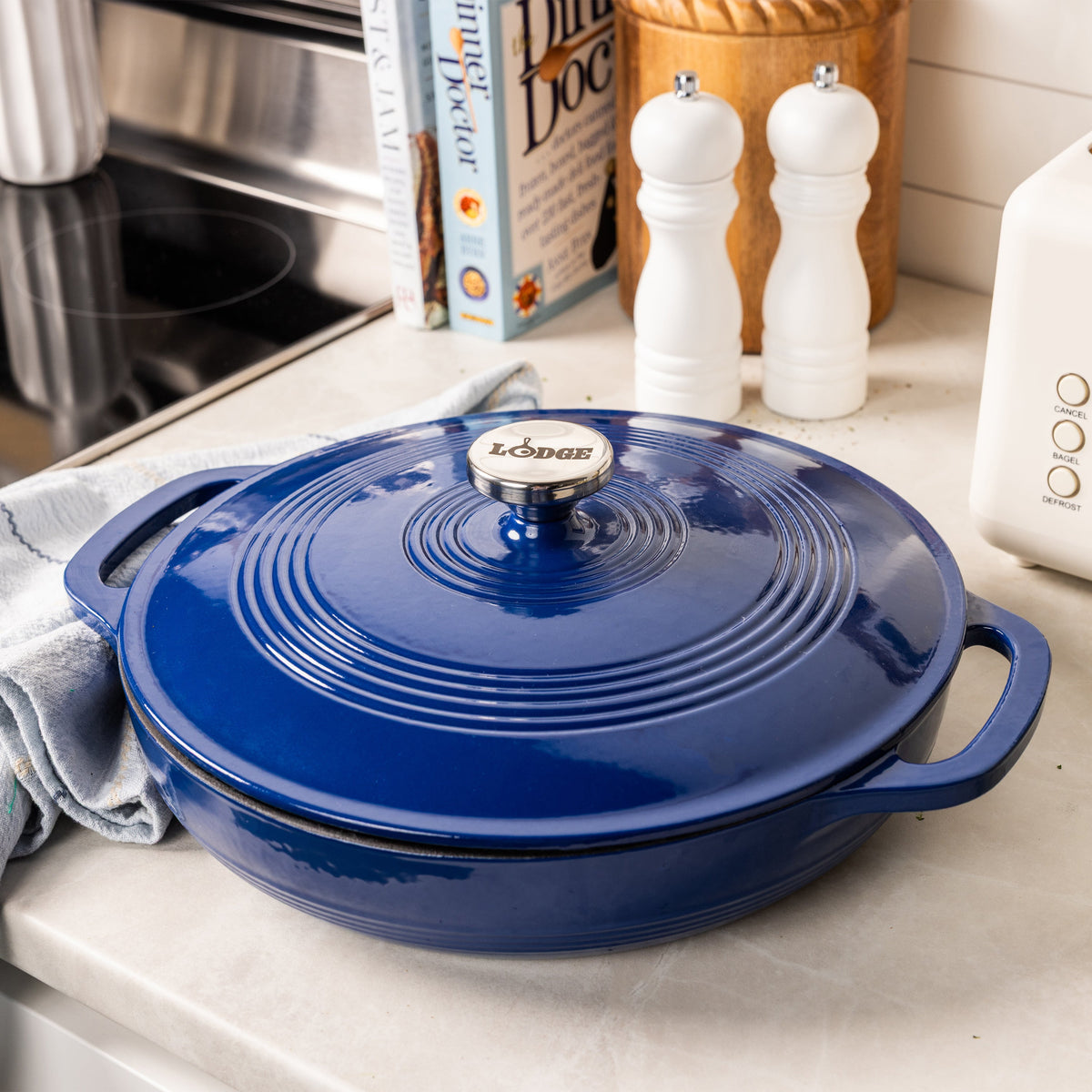 A blue enameled cast iron braiser with a lid, shown on a kitchen counter with a cloth, salt and pepper mills, and cookbooks in the background.