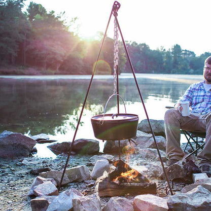 Tripod being used to hold dutch oven over fire while cooking near lake.