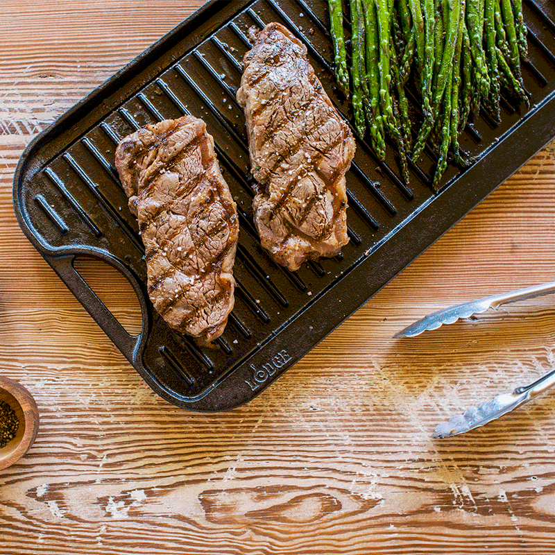 Grilled steaks and asparagus on a cast iron grill pan on a wooden surface.