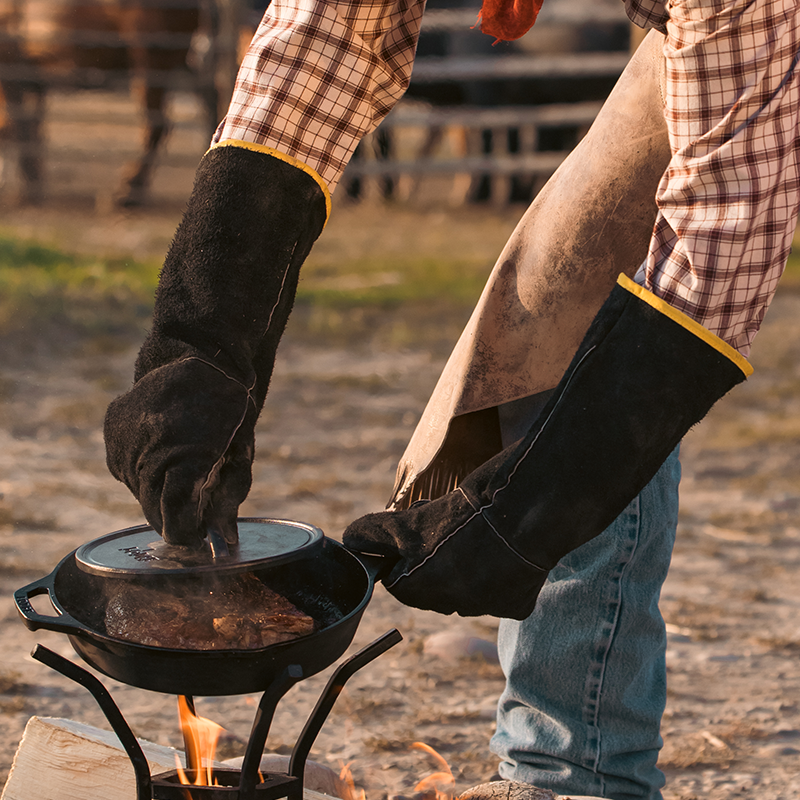 Someone using leather cloves while cooking with a skillet and cast iron grill press over a fire.