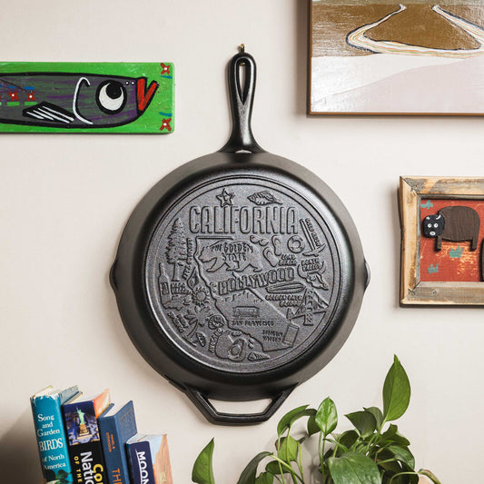 Black cast iron skillet with 'California' engraving on a wall with books and a plant below.