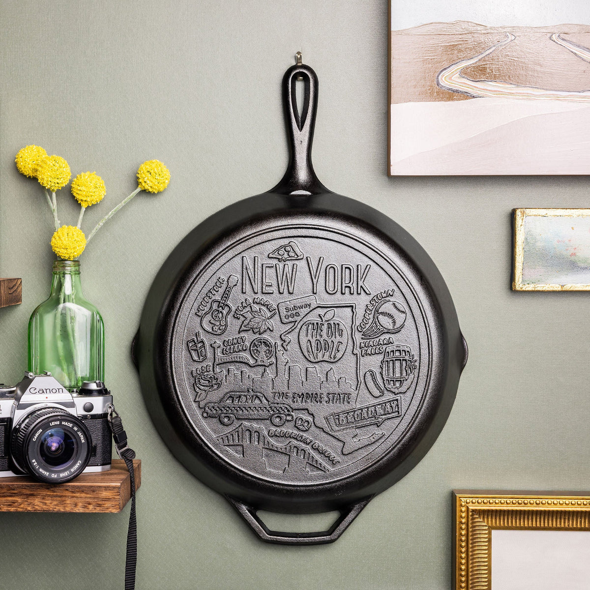 Decorative state cast iron skillet hung a wall with a camera and flowers below.