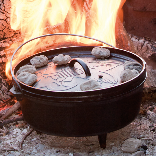 Dutch oven cooking food near camp fire, with hot coals on lid.