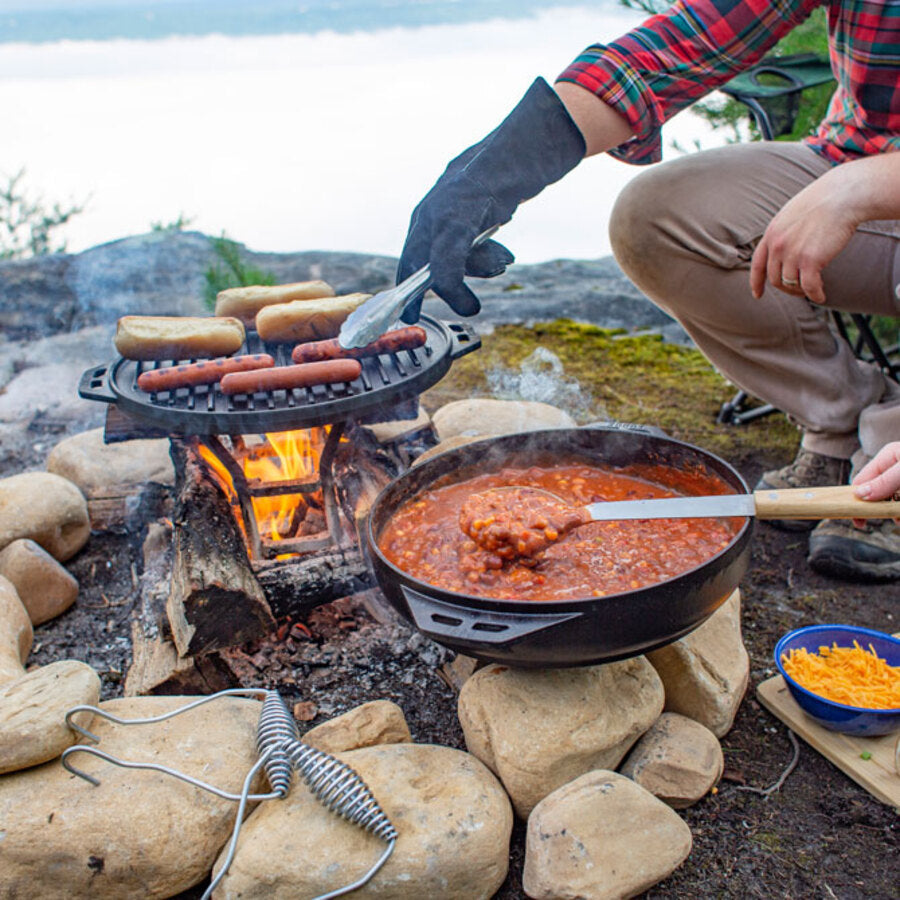 Someone turning hot dogs on grill pan over the fire while someone else stirs chili in wok portion nearby.