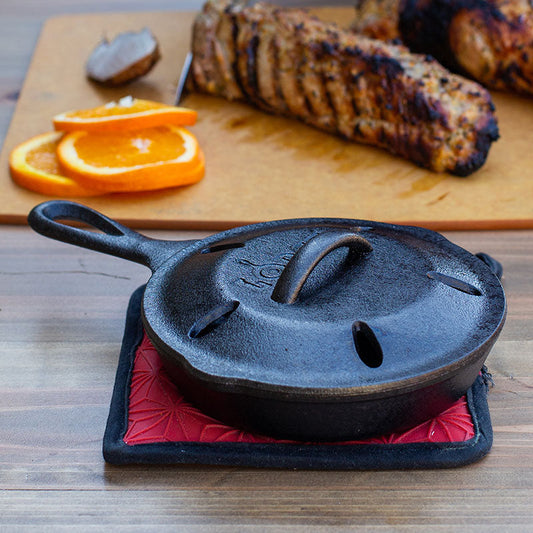 A black cast iron smoker skillet on our silicone and fabric potholder, placed on a wooden surface next to some grilled food and an orange slice.