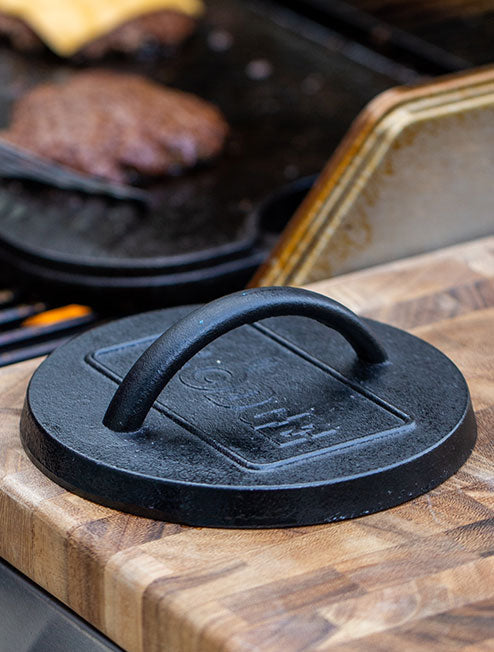 A press sitting next to grill while someone flips a smash burger.