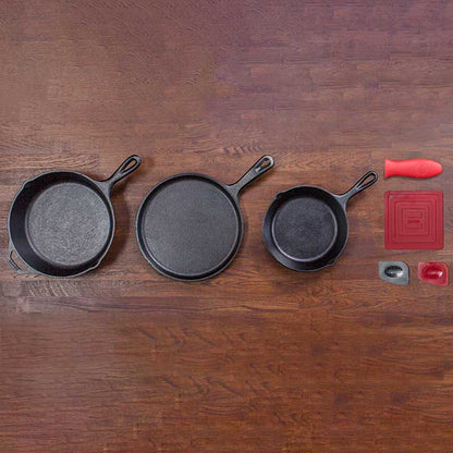 A set of three black cast iron skillets of different sizes placed on a wooden surface, accompanied by a red spatula, a small amount of oil, and a silicone mat.