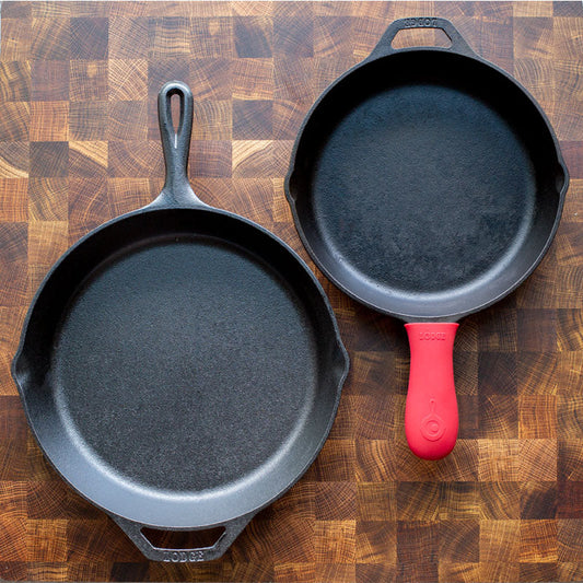 Two black cast iron skillets of different sizes placed on a wooden surface, one with a red silicone handle holder.