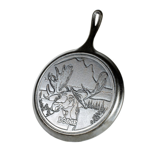 A round cast iron griddle with a moose and mountain landscape design and the word 'LODGE' on it.