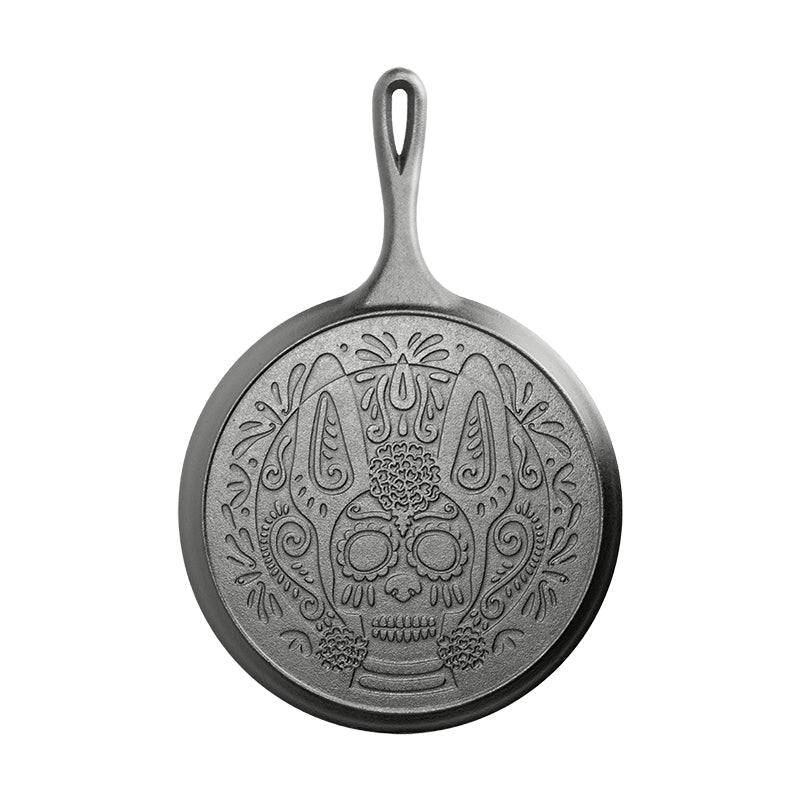 The back of a 10.5 inch round seasoned cast iron griddle with a handle, featuring a sugar skull design resembling a Xolo dog.