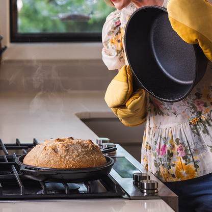 Person wearing yellow oven mitts lifting the deep skillet piece off of skillet piece revealing a freshly baked loaf of bread on a stove.