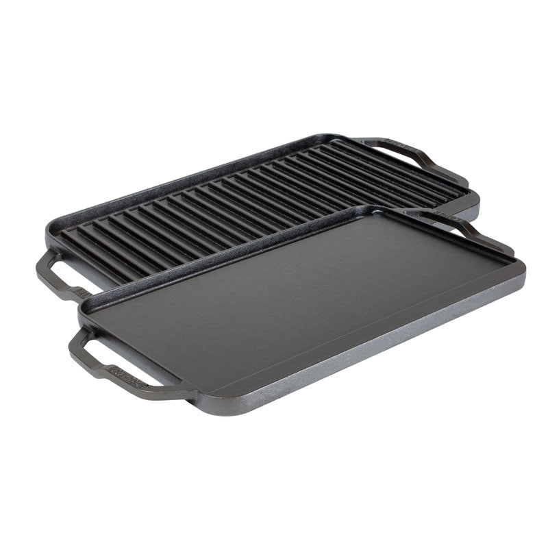 A reversible grill/griddle with one side featuring grooves for grilling and the other side flat, both sides Seasoned with a black seasoning, with lifted handles on the sides.
