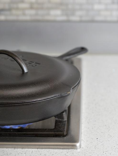 A black cast iron lid placed on top of a cast iron skillet on a stovetop, indicating use for roasting, braising, and simmering.