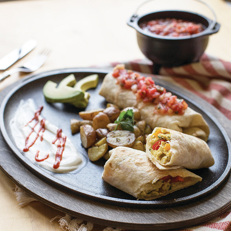 Breakfast burritos and potatoes present on serving griddle with wood underliner.