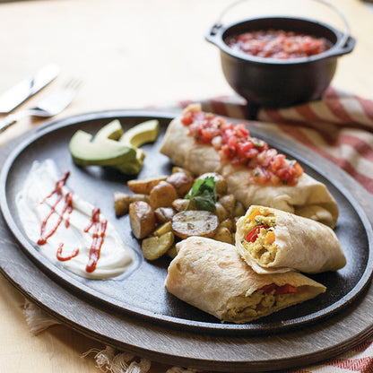 Breakfast burritos and potatoes present on serving griddle with wood underliner.