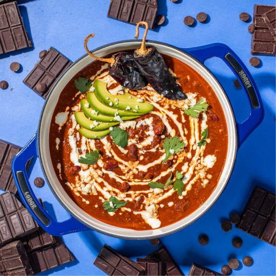 Blue dutch oven with fresh chocolate chili on blue background surrounded by pieces of chocolate.