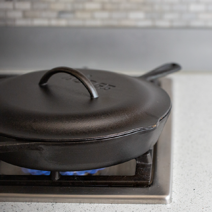 A black cast iron lid placed on top of a cast iron skillet on a stovetop, indicating use for roasting, braising, and simmering.