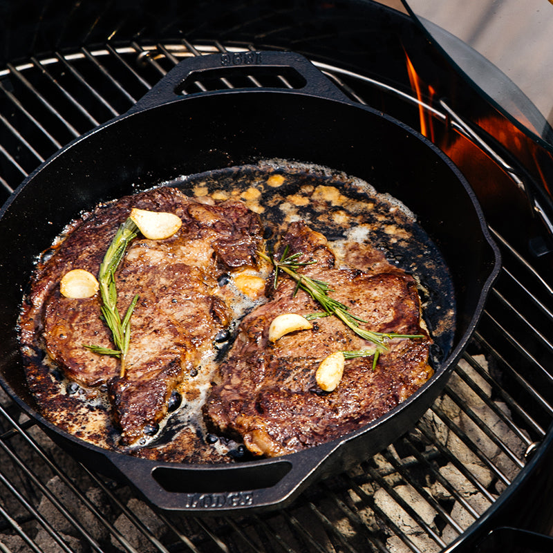 Seared steak with rosemary and garlic being made in pan over charcoal grill.